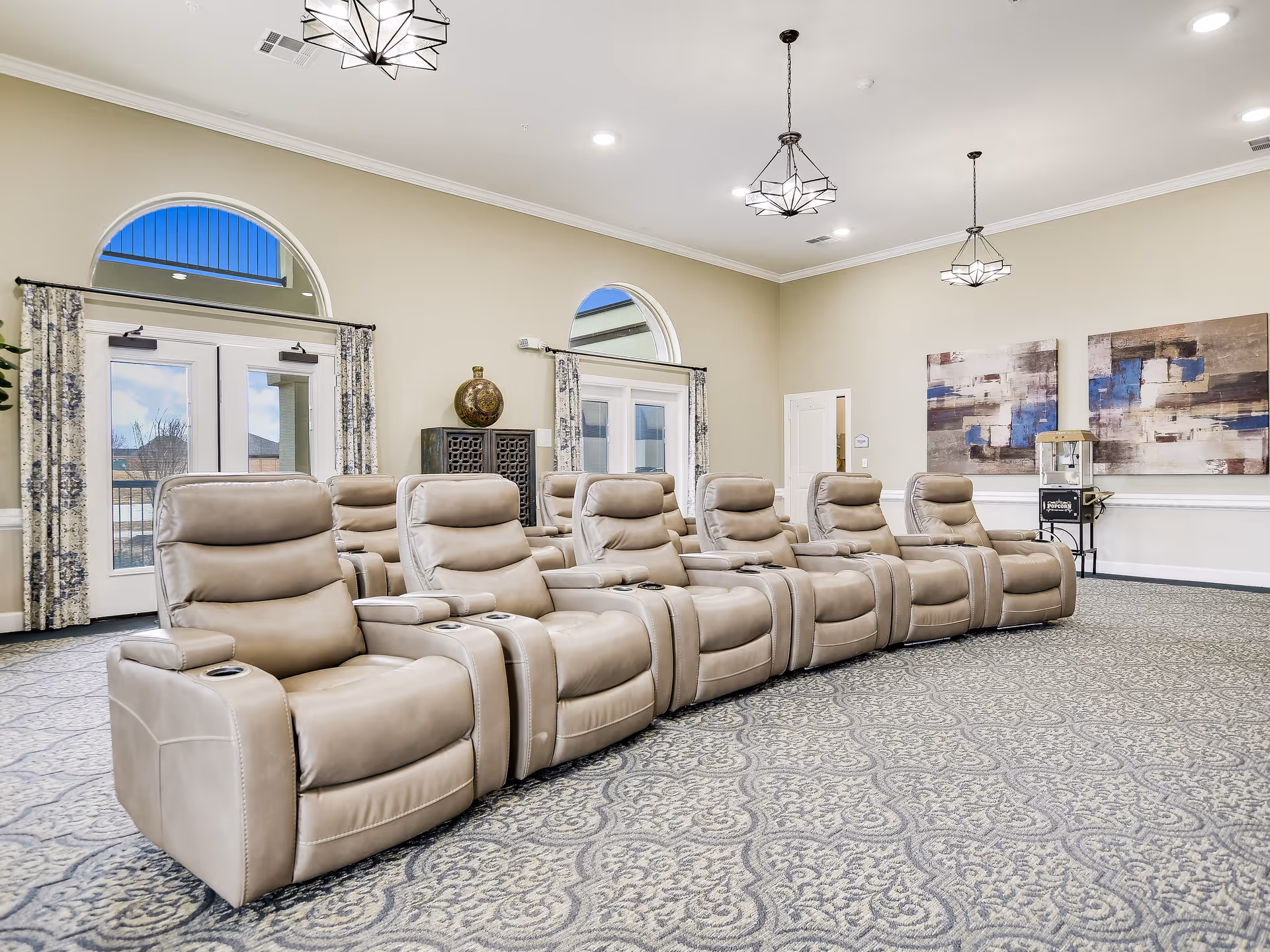 A row of beige reclining theater chairs arranged in a spacious communal media room with patterned carpet, pendant lights, and abstract wall art.