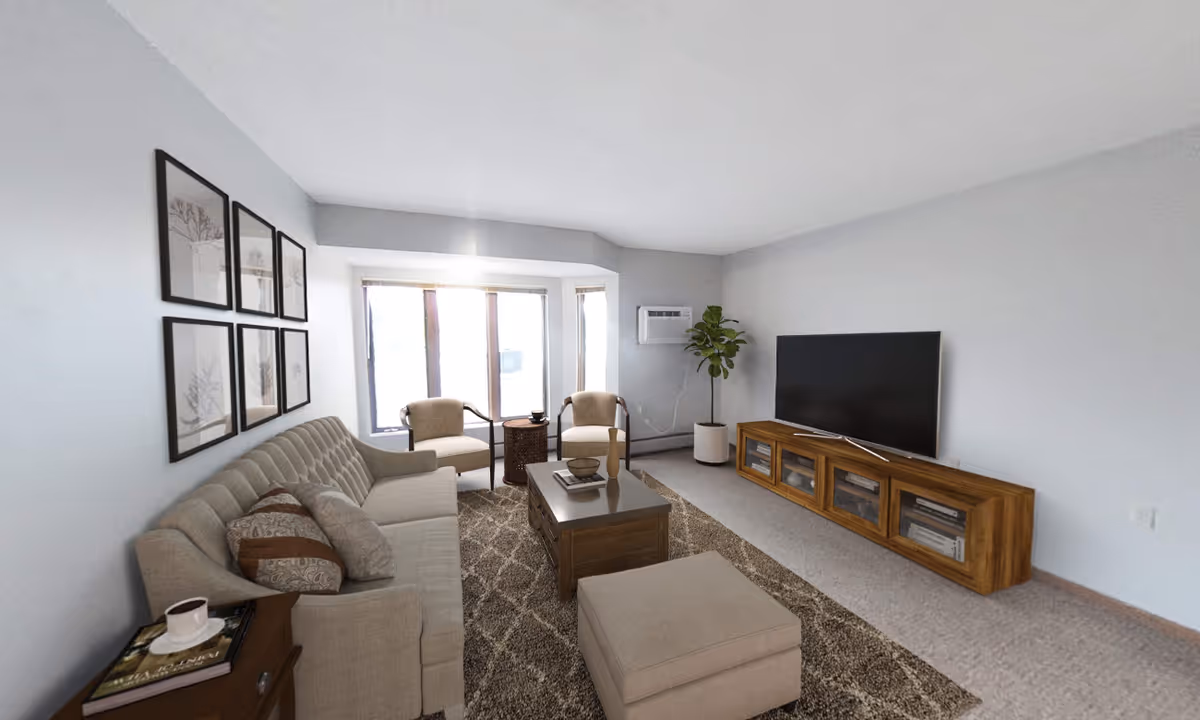 A cozy living room with a beige sofa adorned with patterned pillows, two armchairs near a window, a wooden coffee table, a matching ottoman, a large flat-screen TV on a wooden TV stand, a potted plant in the corner, and framed artwork on the wall.