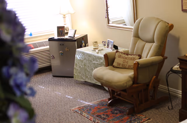A cozy sitting area with a cushioned glider chair bearing a 'Grandmothers give the best hugs' pillow, a small table with photos and lamp, and a mini fridge near a window.