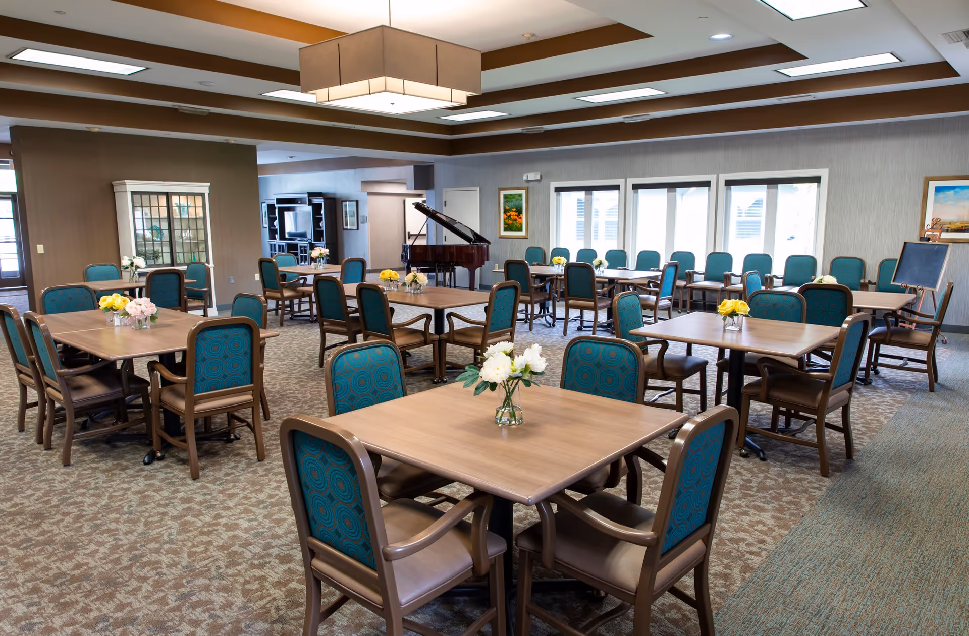 A spacious dining room with multiple square wooden tables surrounded by teal cushioned chairs. Each table has a small vase with flowers. The room features a patterned carpet, large windows letting in natural light, a grand piano in the background, and framed artwork on the walls.
