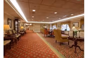Spacious carpeted common sitting area with upholstered chairs, side tables, lamps, and framed artwork in a senior living facility.