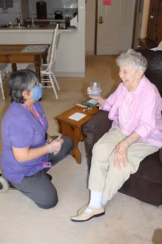 An elderly woman sitting on a couch holding a glass of water, smiling and interacting with a caregiver who is kneeling beside her wearing a purple uniform and a face mask. The setting appears to be a living room with a small wooden table between them and a kitchen visible in the background.