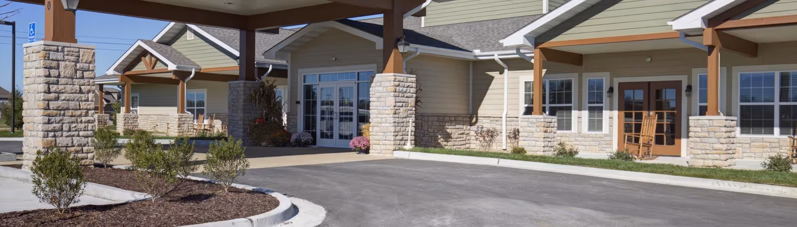 Entrance area of Oak Pointe of Kearney senior living facility showing a covered drop-off zone with stone pillars, a paved driveway, landscaped bushes, and the building's exterior with beige siding and multiple windows.