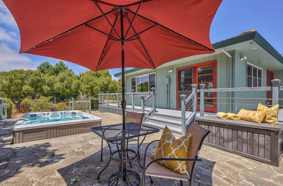 Outdoor patio area with a large red umbrella over a black metal table and chairs with yellow patterned cushions. There is a hot tub on the stone patio and a light green building with red-framed windows and doors in the background. A bench with yellow pillows, one with the word 'RELAX', is also visible.
