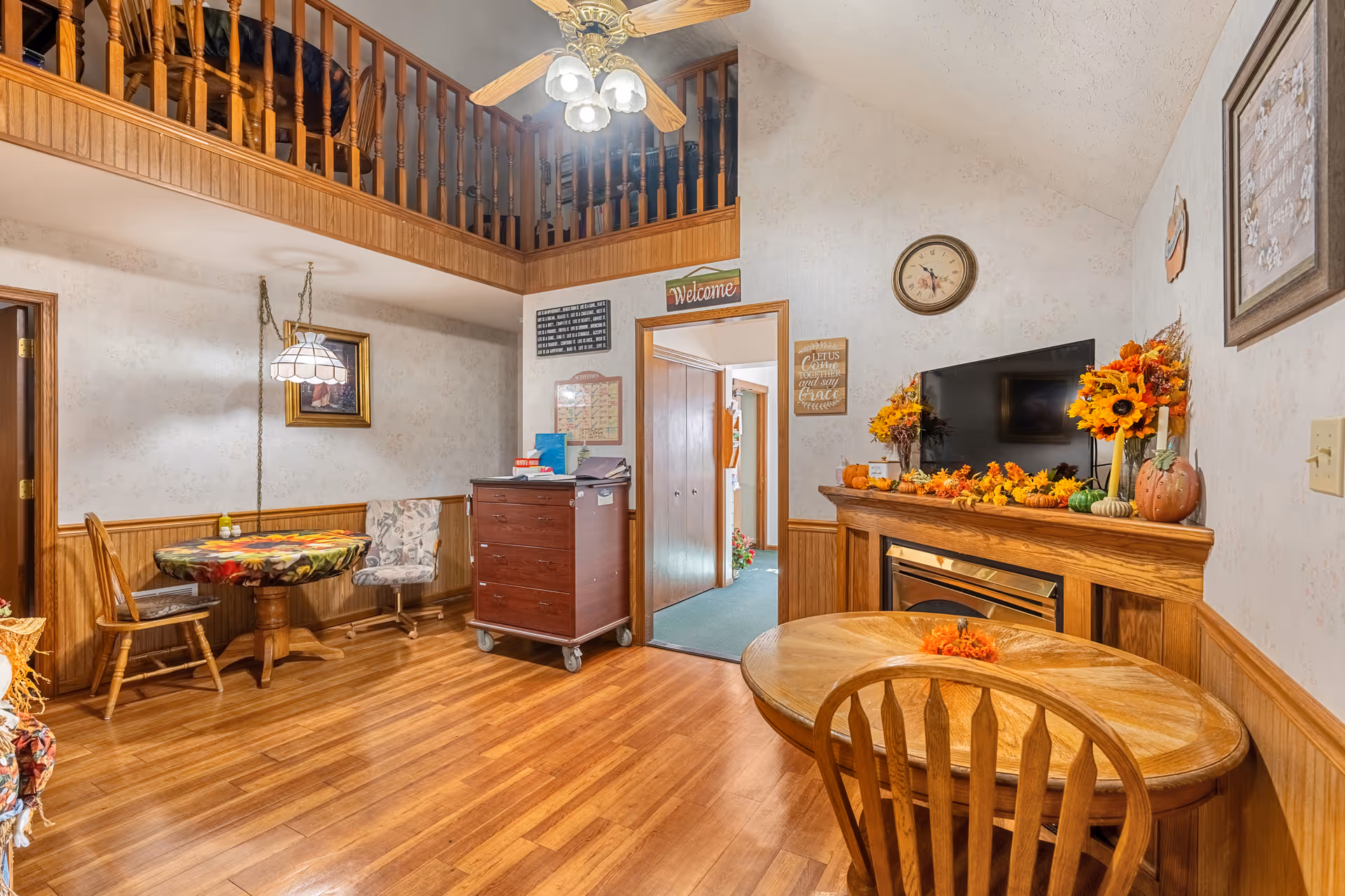 Interior view of a cozy living area in Countryside Home featuring wooden floors, a round wooden table with chairs, a fireplace decorated with autumn-themed flowers and pumpkins, a wall-mounted TV above the fireplace, and a ceiling fan with lights. The room has a high ceiling with a wooden railing balcony above and a small dining nook with a table covered by a floral tablecloth and two chairs. Various framed pictures and decorative signs adorn the walls.