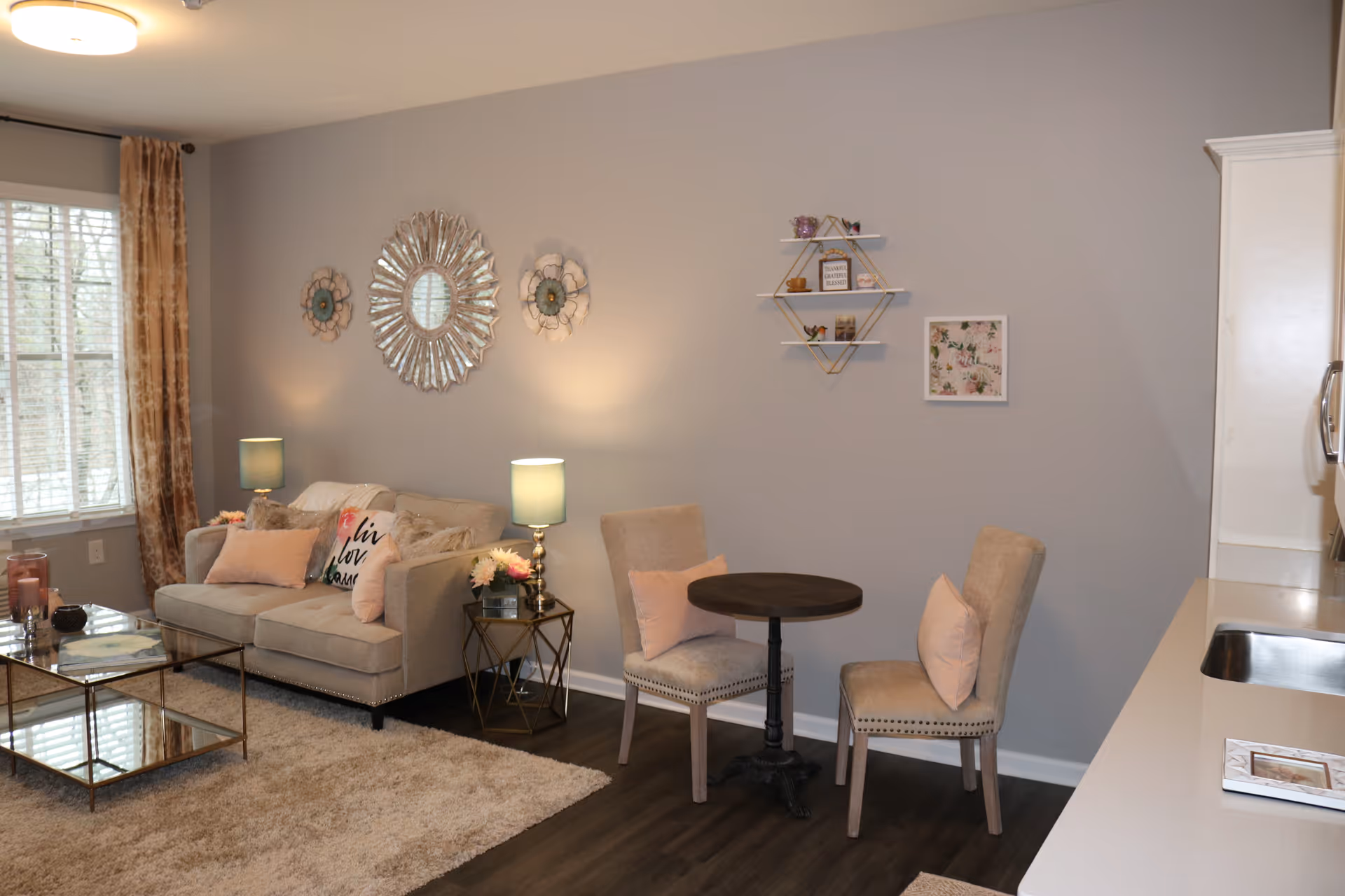 A cozy living room area with a beige sofa adorned with decorative pillows, two side tables with lamps, a round small dining table with two upholstered chairs, wall decorations including a large sunburst mirror and small shelves, a window with curtains, and a kitchen counter with a sink visible on the right side.