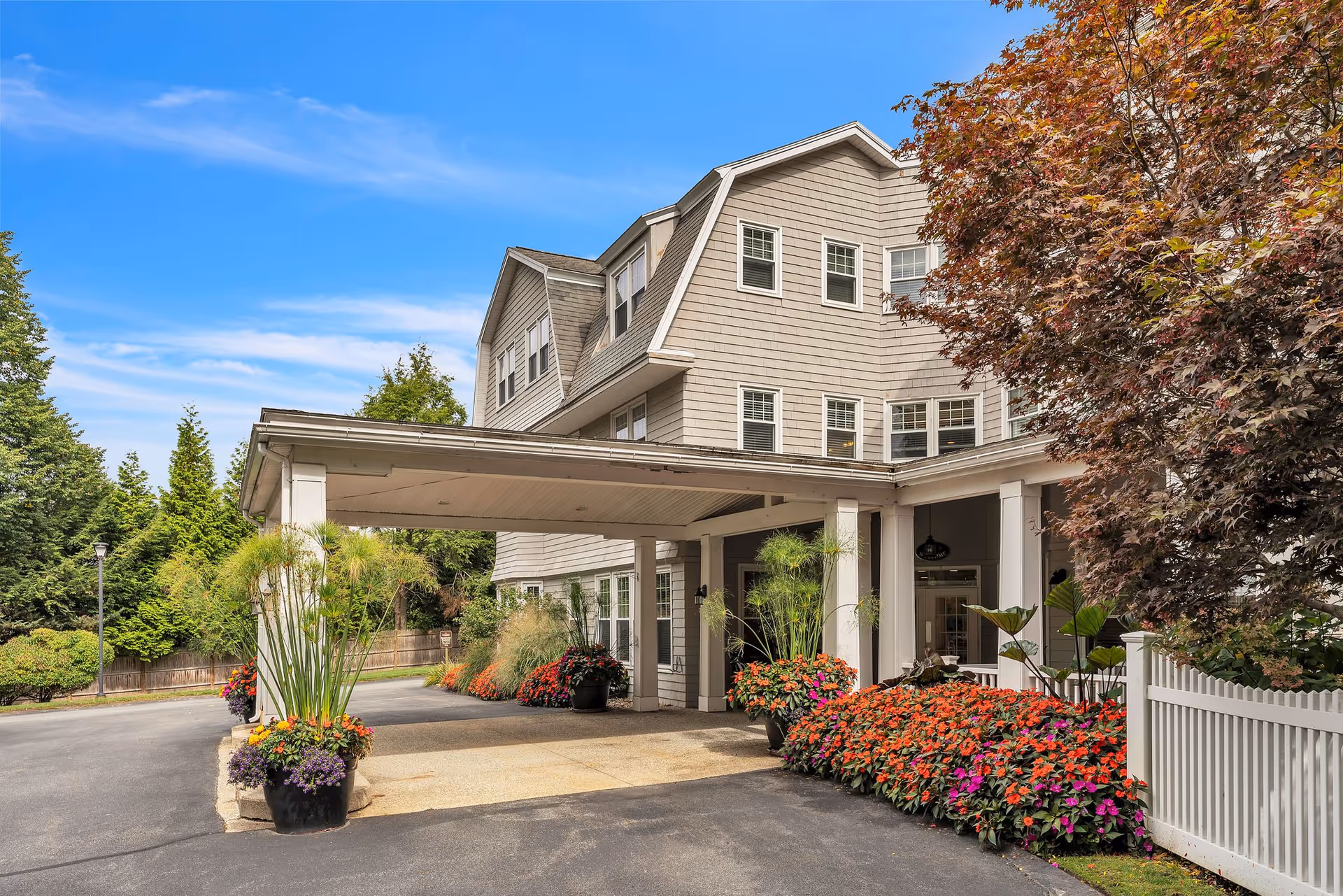 Front entrance of a multi-story senior living building with a covered porte-cochere, flowering planters, and surrounding trees.