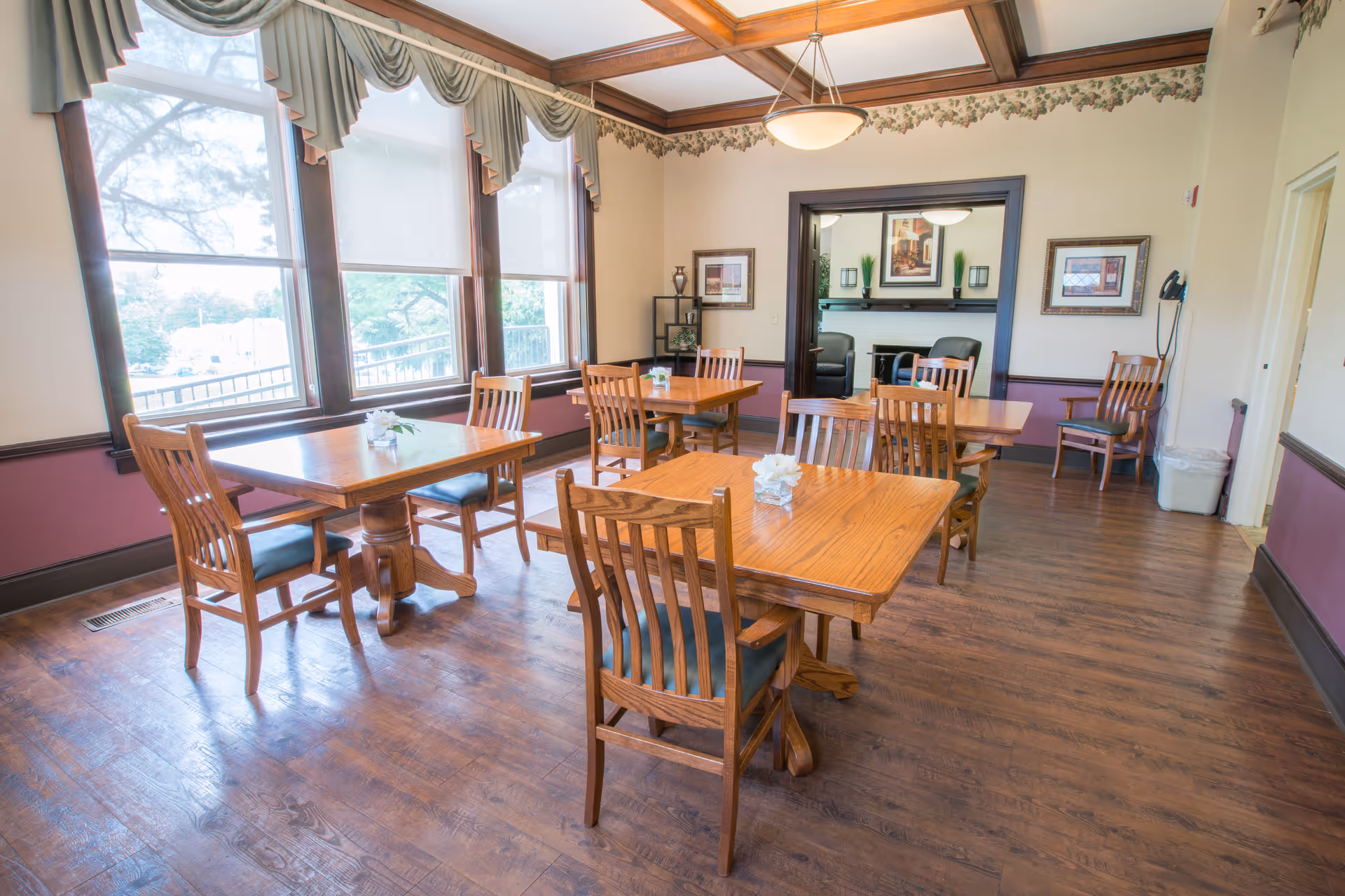 A bright dining room with wooden tables and chairs arranged neatly. Large windows with valance curtains allow natural light to fill the room. The walls are decorated with framed pictures and a floral border near the ceiling. The floor is wooden, and there is a doorway leading to another room with additional seating.