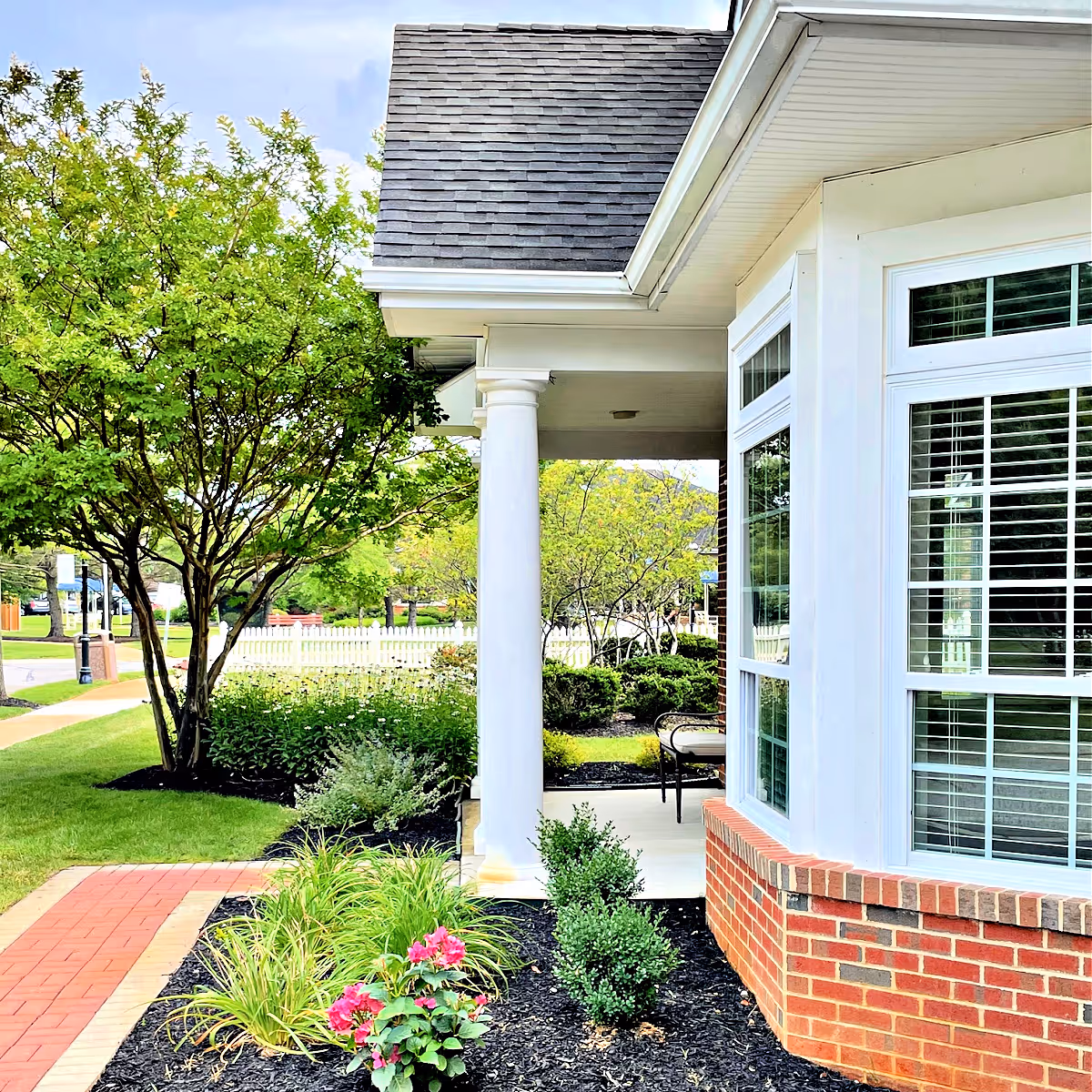 View of the exterior corner of a building with white framed windows and a brick base, a covered porch supported by a white column, surrounded by landscaped greenery including bushes, flowering plants, and a tree, with a red brick walkway leading alongside the building.