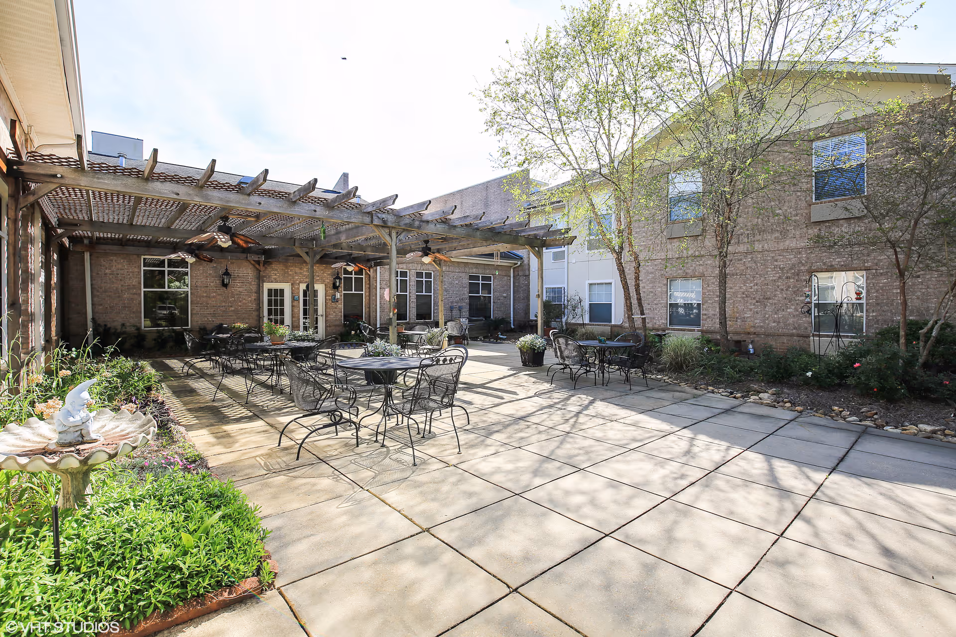 Outdoor patio area at The Waterford on Highland Colony featuring several black metal tables and chairs under a wooden pergola with ceiling fans. The patio is surrounded by a brick building with windows and doors, and there are landscaped garden beds with greenery and a small birdbath statue.