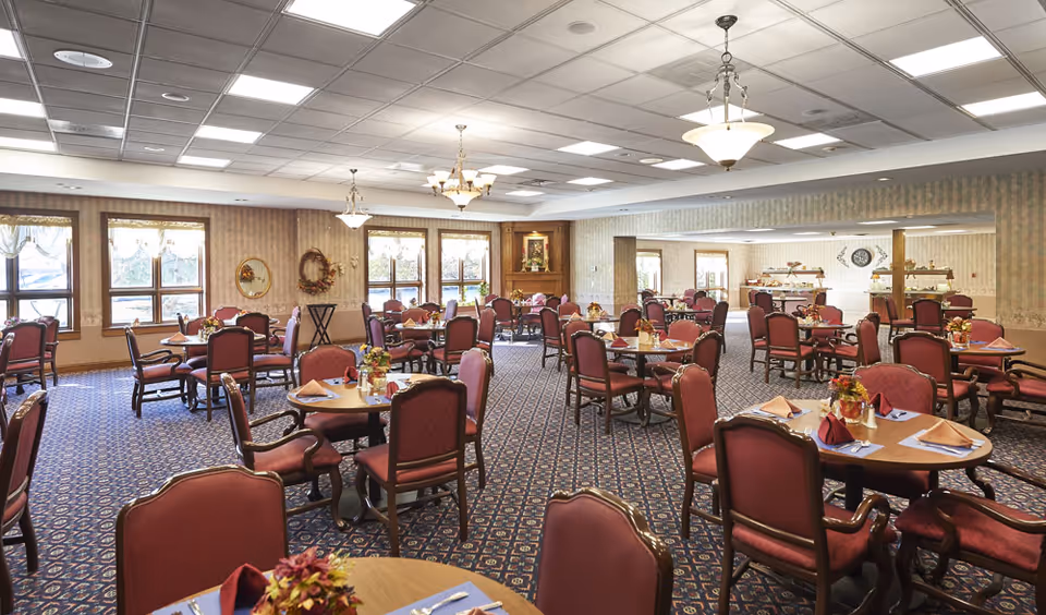 A spacious dining room with multiple round tables set with napkins, silverware, and small floral centerpieces. The room features large windows with curtains, patterned carpet, and chandeliers hanging from the ceiling. The walls are decorated with wreaths and framed artwork.