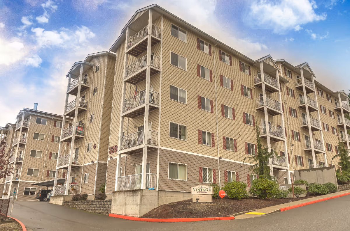 Exterior view of a multi-story senior living facility building with beige siding and red shutters. The building has multiple balconies and is situated on a sloped driveway with landscaping and a sign that reads Vintage at Silverdale.