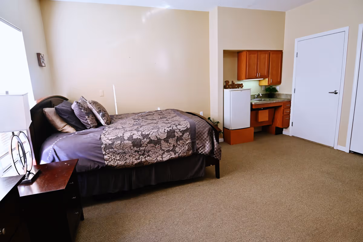 A bedroom in a senior living facility featuring a bed with patterned bedding and multiple pillows, a nightstand with a lamp, a small kitchenette area with a mini refrigerator, sink, and wooden cabinets, beige walls, and carpeted floor.