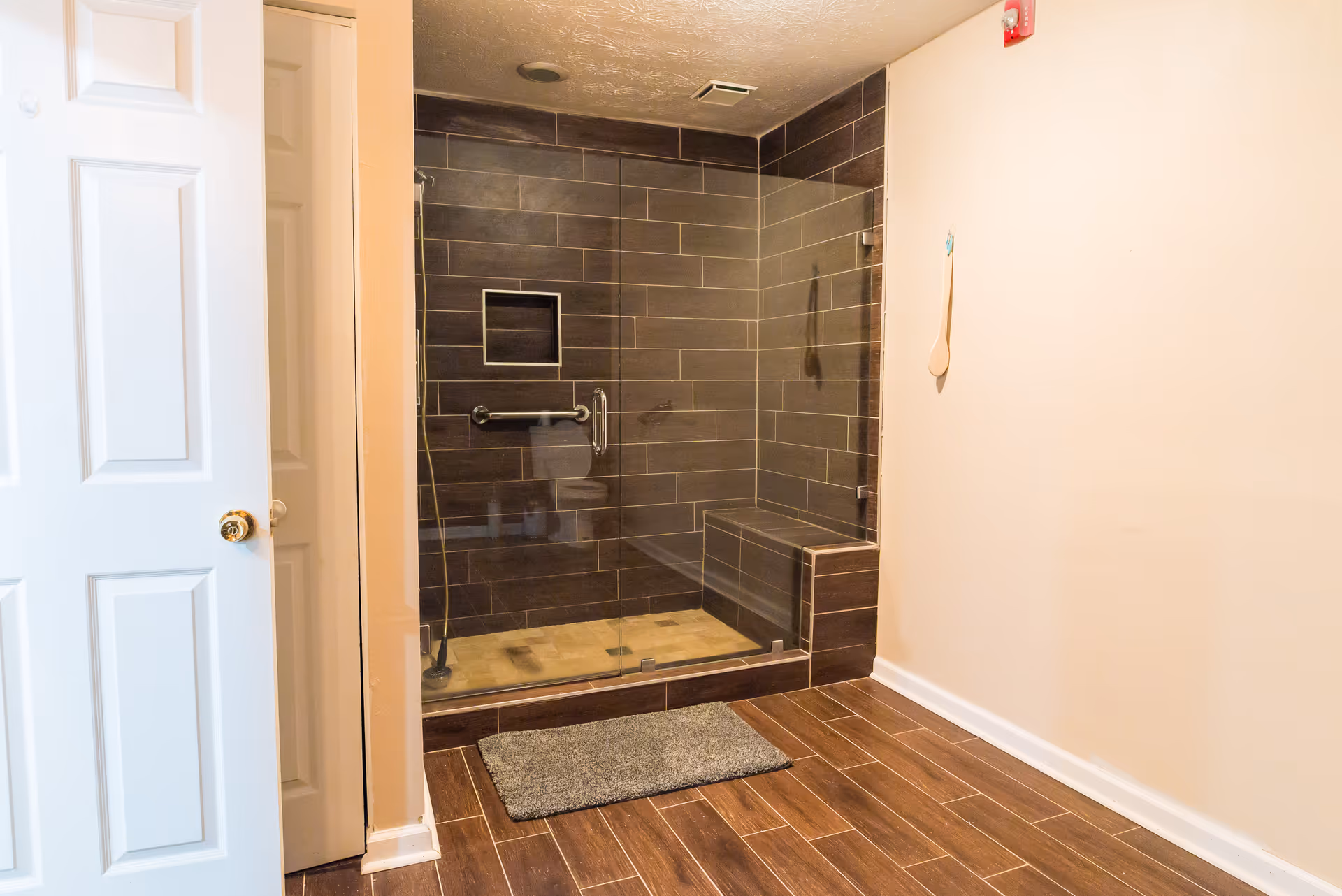 Glass-enclosed tiled walk-in shower with a built-in bench and grab bar, wood-look floor, and an open white door.