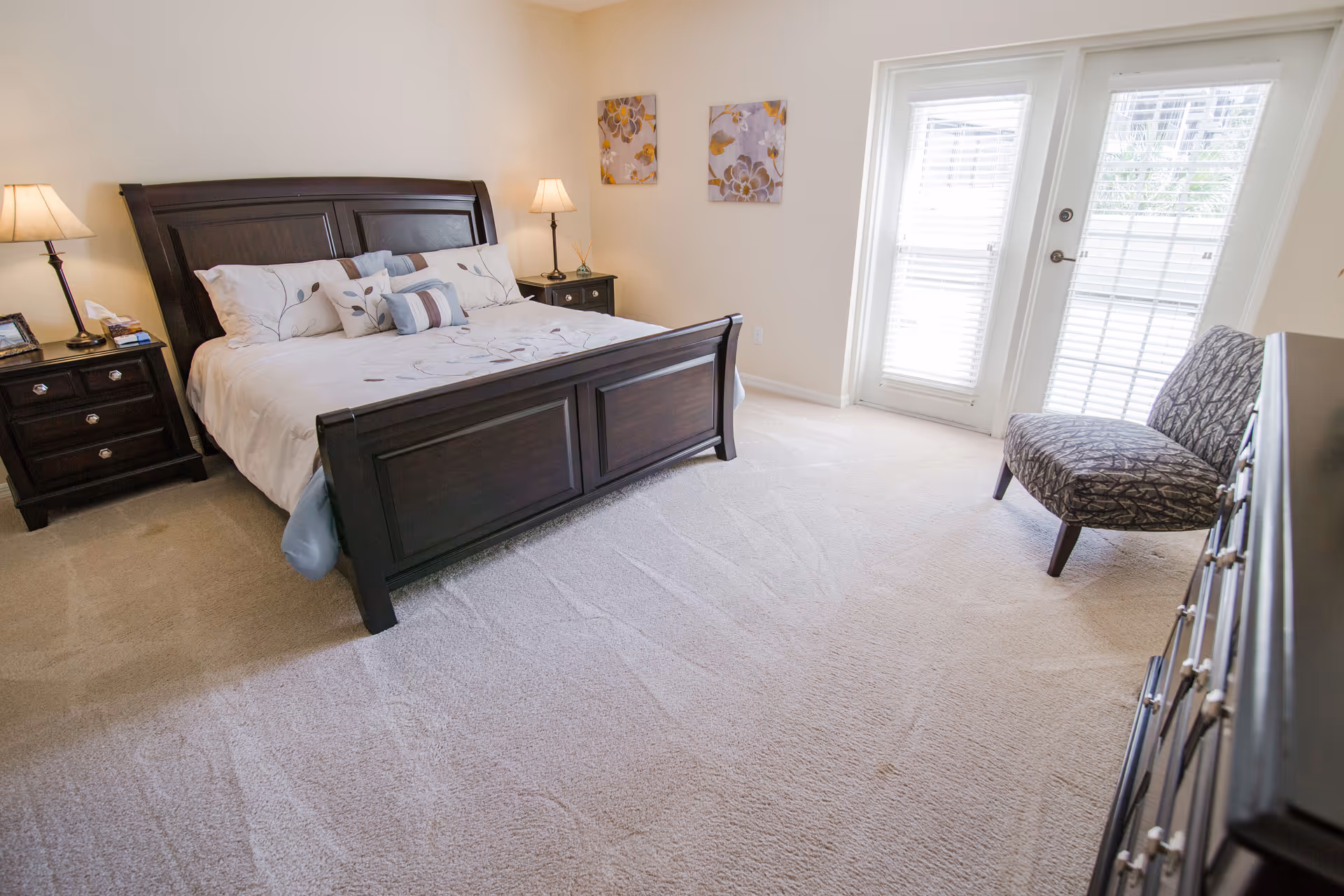 A bright bedroom with a large dark wooden bed frame and white bedding with floral patterns. There are two matching nightstands with lamps on either side of the bed. Two floral paintings hang on the wall above the bed. A patterned chair is placed near a set of glass doors with white blinds, and a dark wooden dresser is visible on the right side of the room. The floor is carpeted in a light beige color.