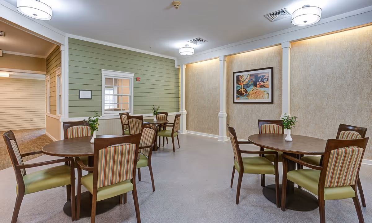 Dining room with round wooden tables, striped and green-upholstered chairs, and framed artwork on the wall.