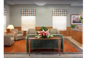 A cozy living room area with a green sofa, two patterned armchairs, a wooden coffee table with a floral arrangement, and a wooden sideboard with a vase of flowers. The room has two windows with patterned valances and a framed picture on the wall.