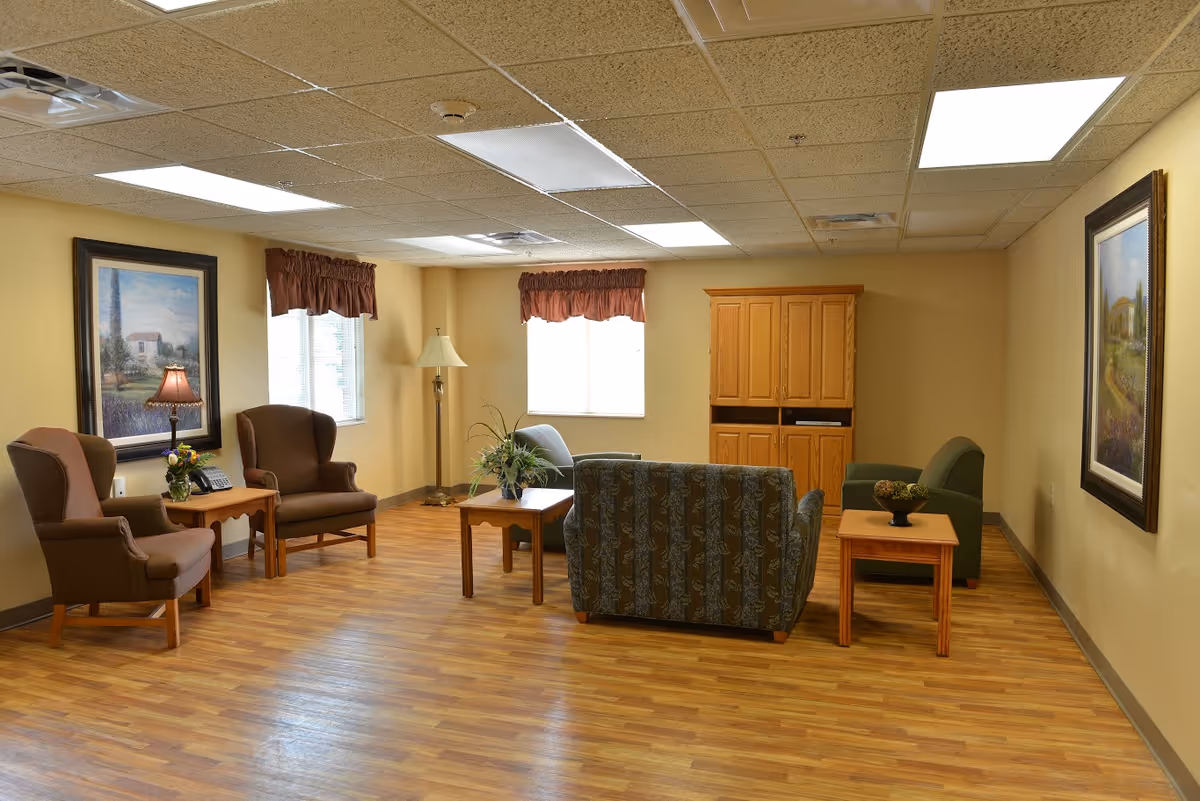 A cozy living room area with wooden flooring, two armchairs, a loveseat, a small sofa, two side tables with plants and a lamp, a wooden cabinet, and framed paintings on the walls. The room is well-lit with ceiling lights and natural light from two windows with brown valances.