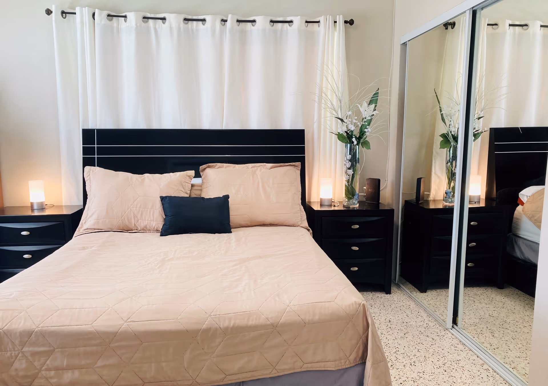 A neatly made bed with beige bedding and three pillows, two beige and one dark blue, against a black headboard. On each side of the bed are black nightstands with drawers, each holding a small lamp. Behind the bed is a white curtain, and to the right is a mirrored sliding closet door reflecting part of the room. A tall vase with white flowers and green leaves is placed on the right nightstand.