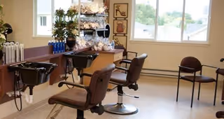 Interior view of a hair salon area with two black salon chairs in front of sinks and mirrors, shelves with hair products and towels, and a window showing an outdoor view.