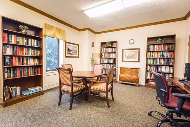 A cozy room with three wooden bookshelves filled with books, a round wooden table surrounded by four striped upholstered chairs, a wooden chest of drawers, a floor lamp, framed artwork on the walls, and a window with a yellow valance. There is also a desk with a computer and an office chair on the right side.