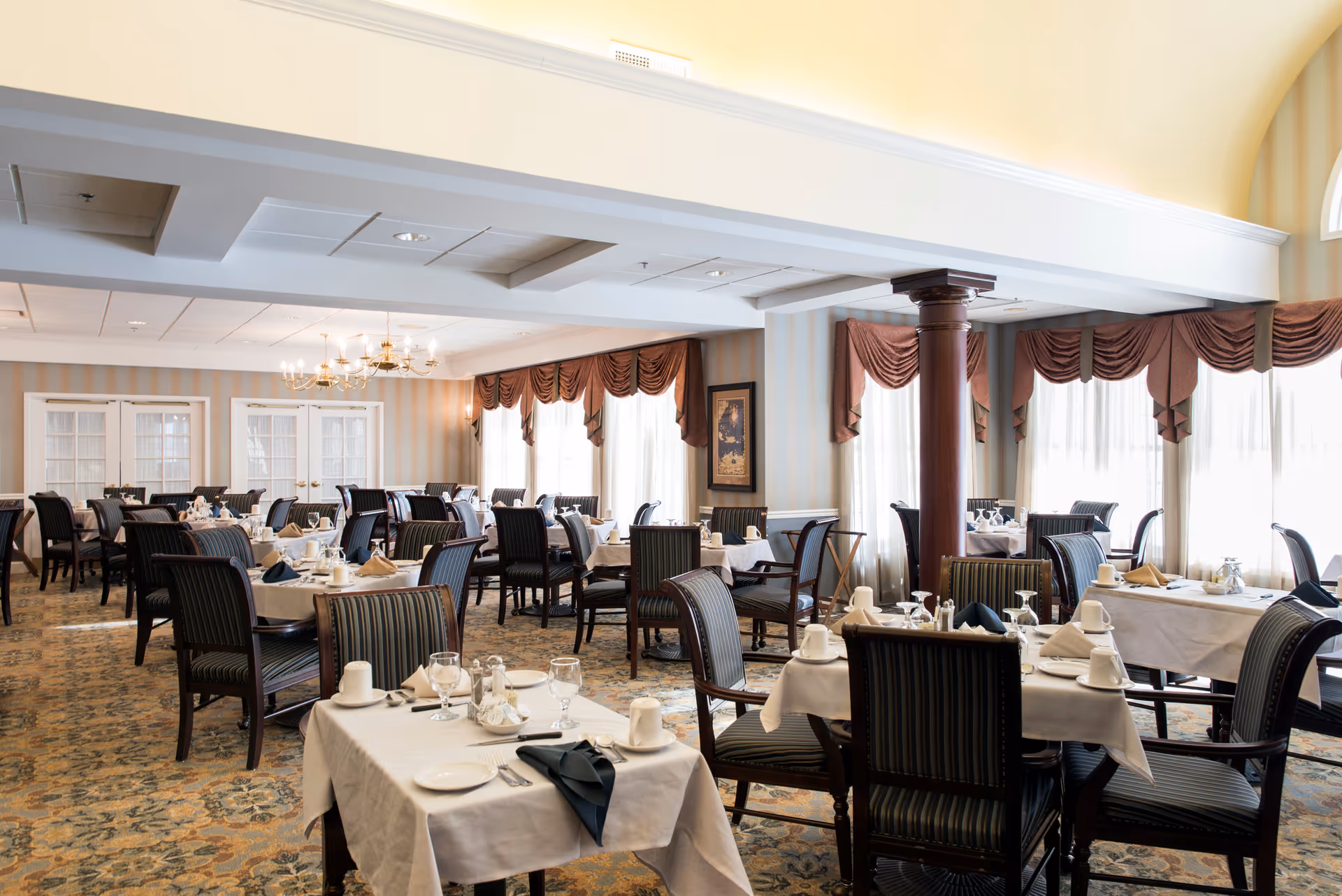 A spacious dining room with multiple tables covered in white tablecloths, set with plates, glasses, napkins, and silverware. The room features large windows with sheer curtains and decorative valances, striped upholstered chairs, a patterned carpet, and a chandelier hanging from the ceiling.