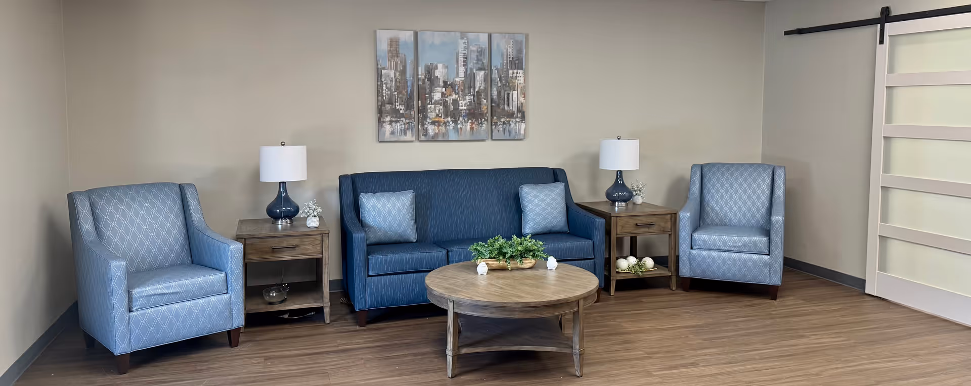 A cozy sitting area with a blue sofa and two matching blue armchairs arranged around a round wooden coffee table with a green plant centerpiece. There are two wooden side tables with blue lamps and decorative items. A three-panel cityscape painting hangs on the beige wall behind the sofa, and a sliding barn door is visible on the right side of the room. The floor is wood laminate.