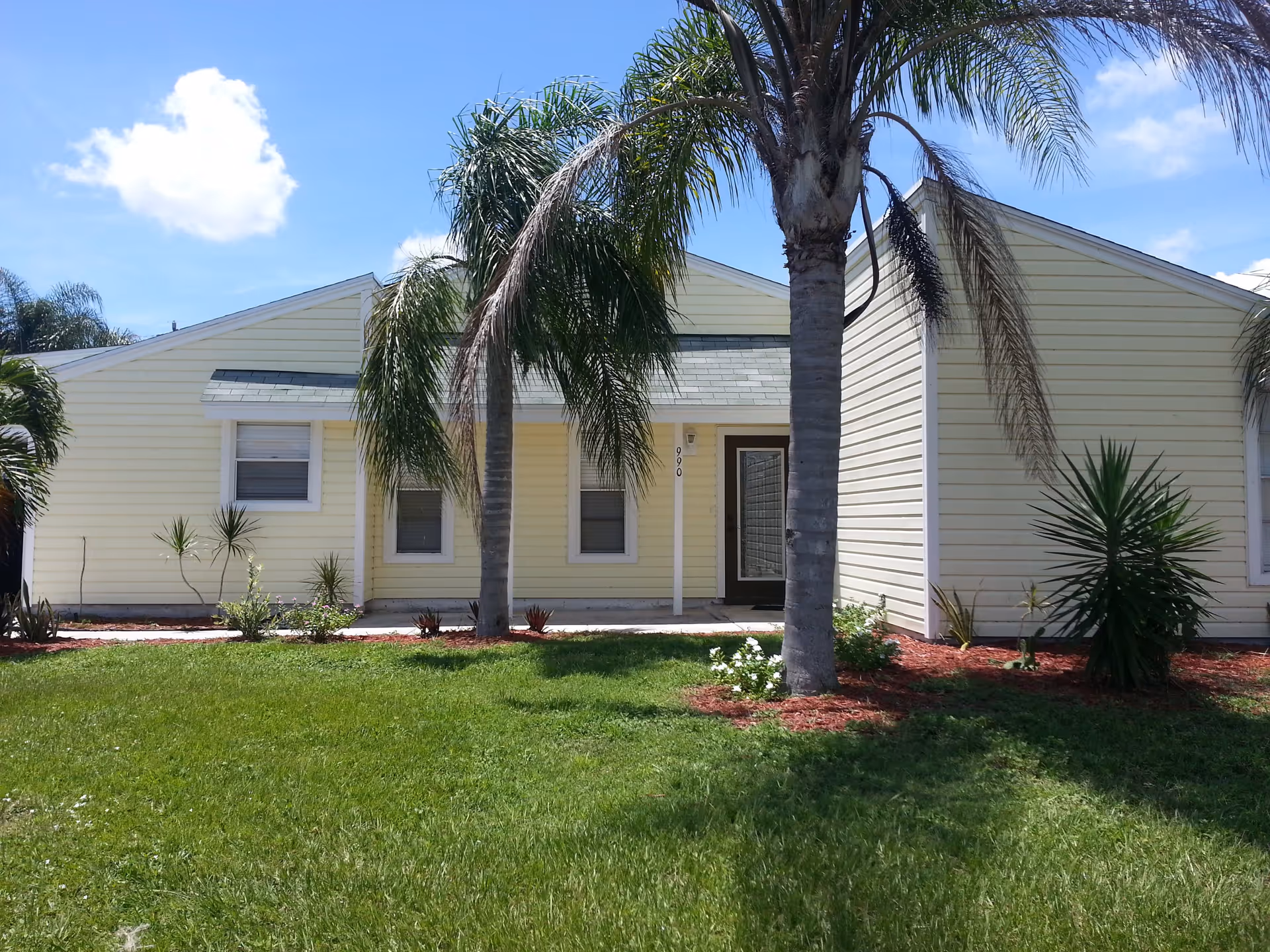 Exterior view of a single-story yellow building with a green lawn and palm trees in front under a blue sky with a few clouds.