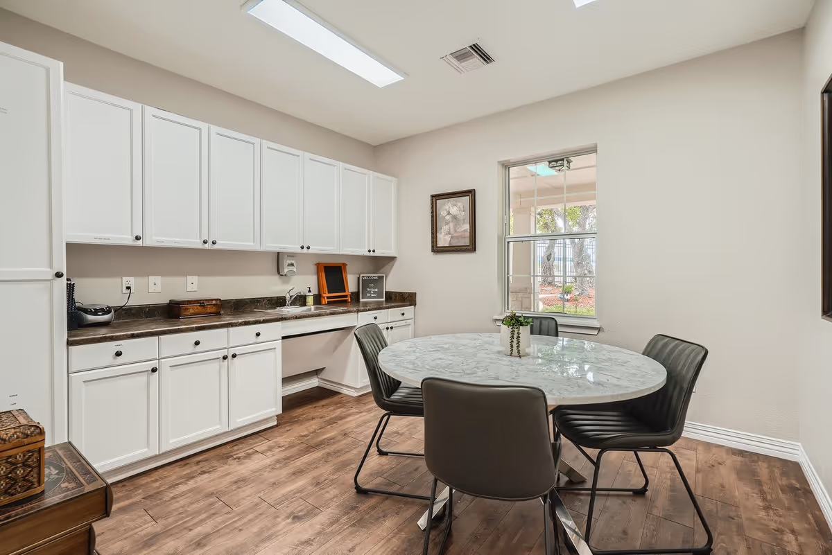 A bright room with white cabinets along one wall and a round marble table surrounded by four modern black chairs. There is a window showing a view of trees outside, a framed picture on the wall, and a small plant on the table. The floor is wood with a warm tone.
