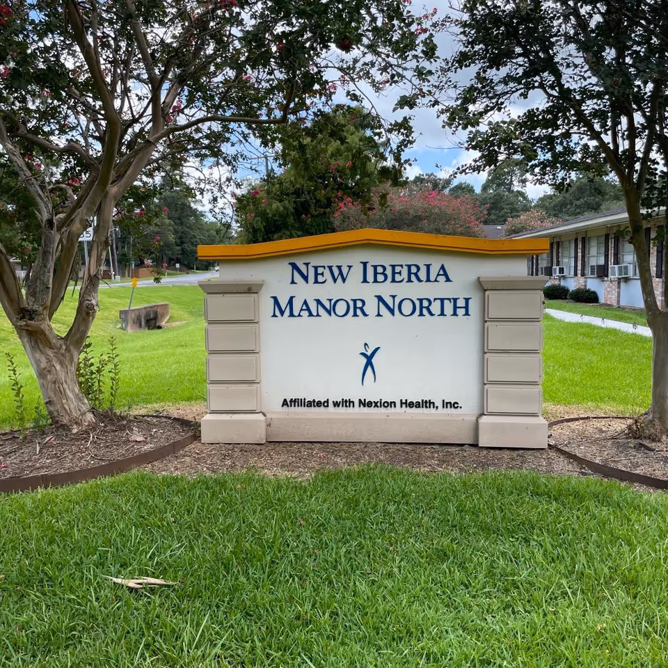 Outdoor view of a sign for New Iberia Manor North, a senior living facility affiliated with Nexion Health, Inc. The sign is white with blue text and a yellow top border, situated on a grassy area with trees and a building in the background.