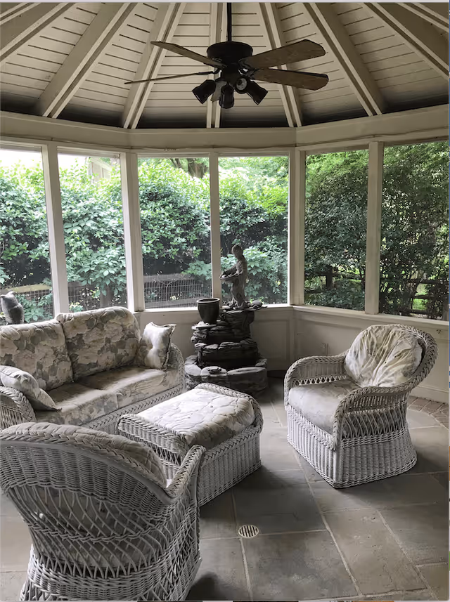A cozy enclosed patio area with white wicker furniture including a sofa, two armchairs, and an ottoman with floral cushions. There is a ceiling fan with lights above, and large windows surround the space showing green bushes and trees outside. A small decorative statue and a potted plant are placed on a stone pedestal in the corner.
