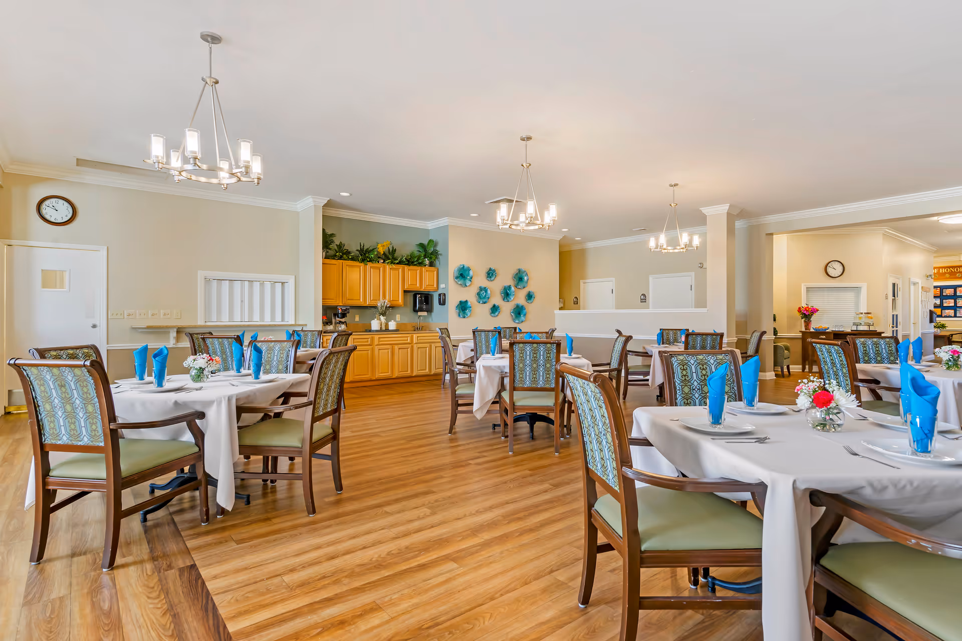 A bright dining room with several round tables covered with white tablecloths, each set with plates, silverware, and blue folded napkins. The room has wooden flooring, wooden chairs with patterned upholstery, modern chandeliers, and a cabinet area with plants and a coffee machine. The walls are decorated with blue floral wall art and there are clocks on the walls.