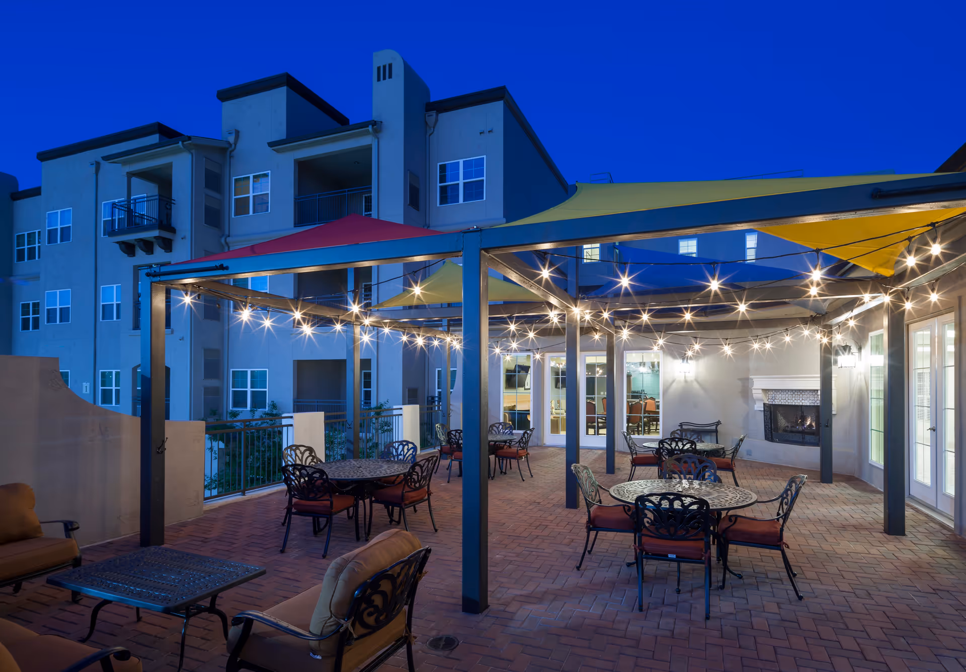 Outdoor patio area at night with multiple round tables and chairs under a metal pergola with colorful shade sails and string lights. The patio has brick flooring and a fireplace on one side. The background shows a multi-story building with balconies and windows.