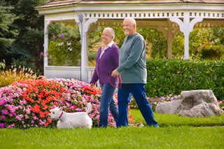 Two older adults walking a small white dog on a green lawn in front of colorful flowerbeds and a white gazebo.