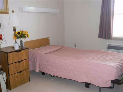Single bed with pink spread, dresser, lamp, sunflowers, and window in bedroom.