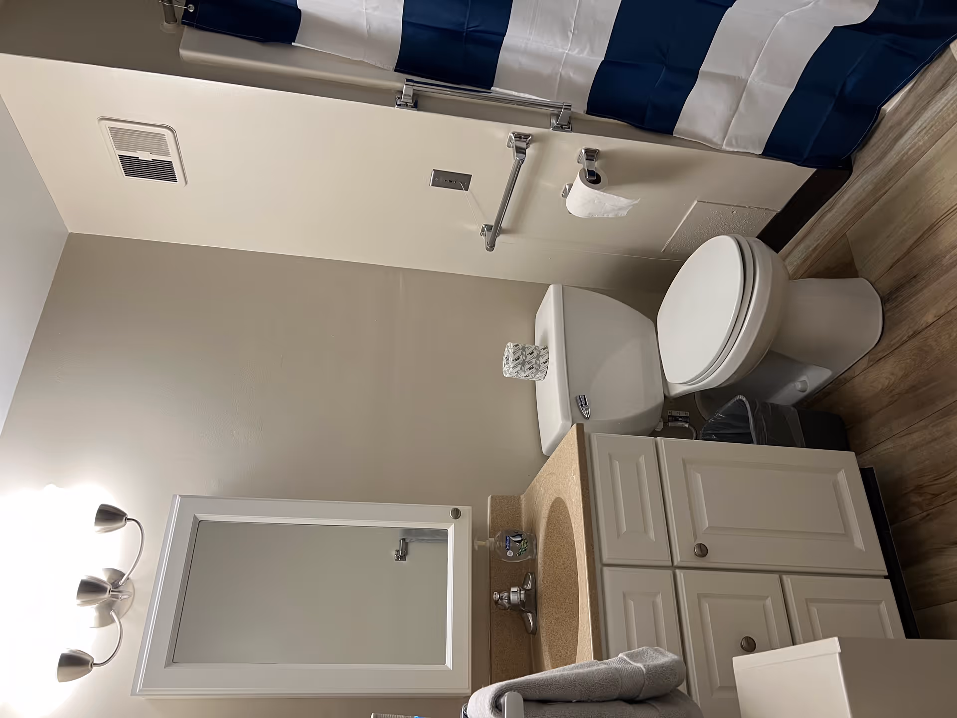 A small bathroom with a white toilet, a beige countertop with a sink, a white cabinet with a mirror above it, a towel hanging on a rack, a trash bin, and a shower with a blue and white striped curtain.