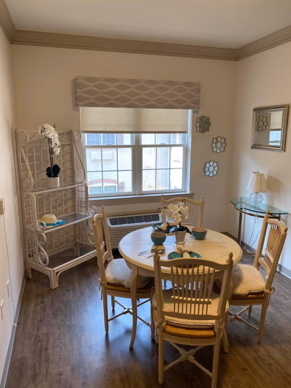 Small dining area with a round table set for four, wooden chairs, a window, shelving unit and side table with a lamp.