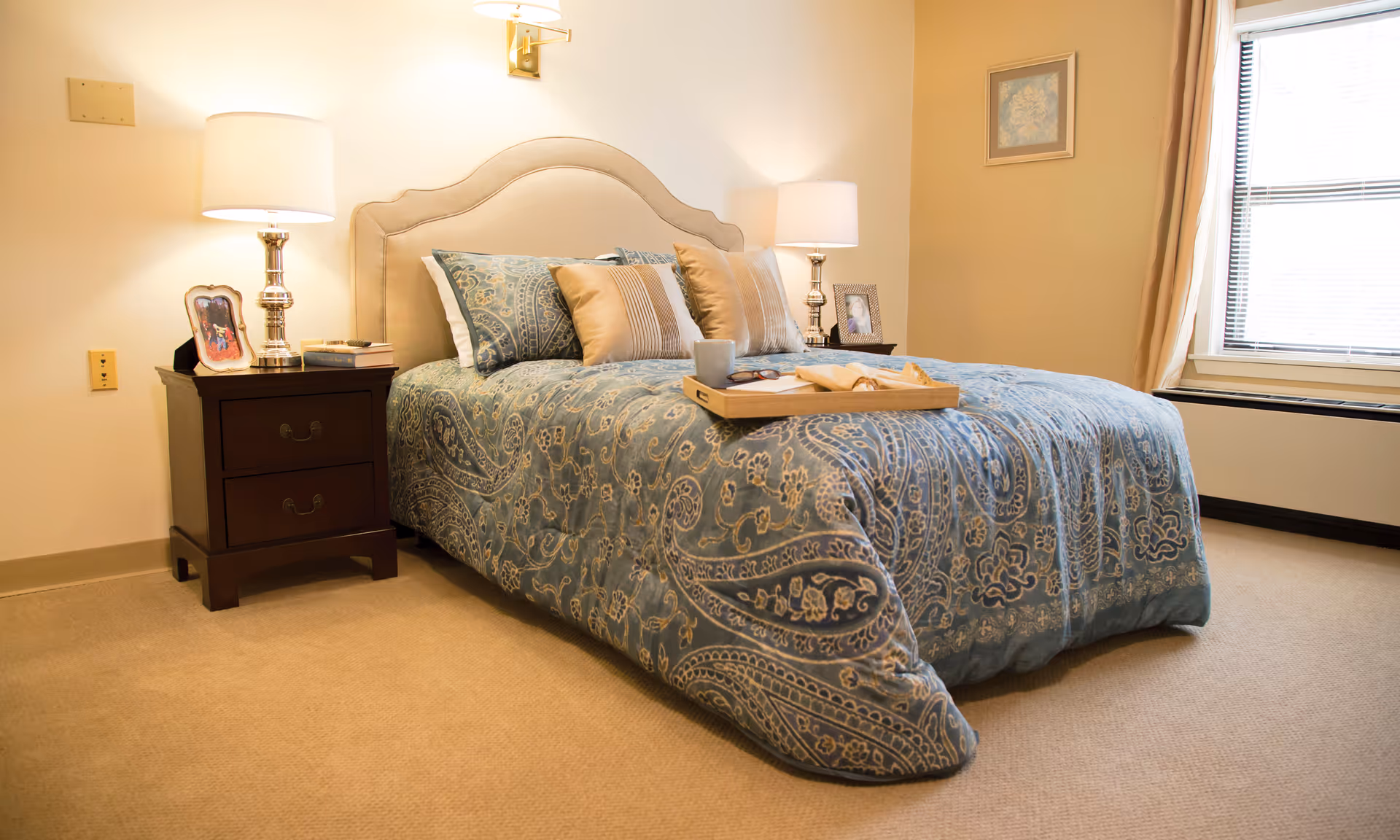 A cozy bedroom with a bed covered in a blue patterned comforter and several pillows. There are two bedside tables with lamps, framed photos, and books. A window with blinds and curtains is on the right side, and a framed picture hangs on the wall.