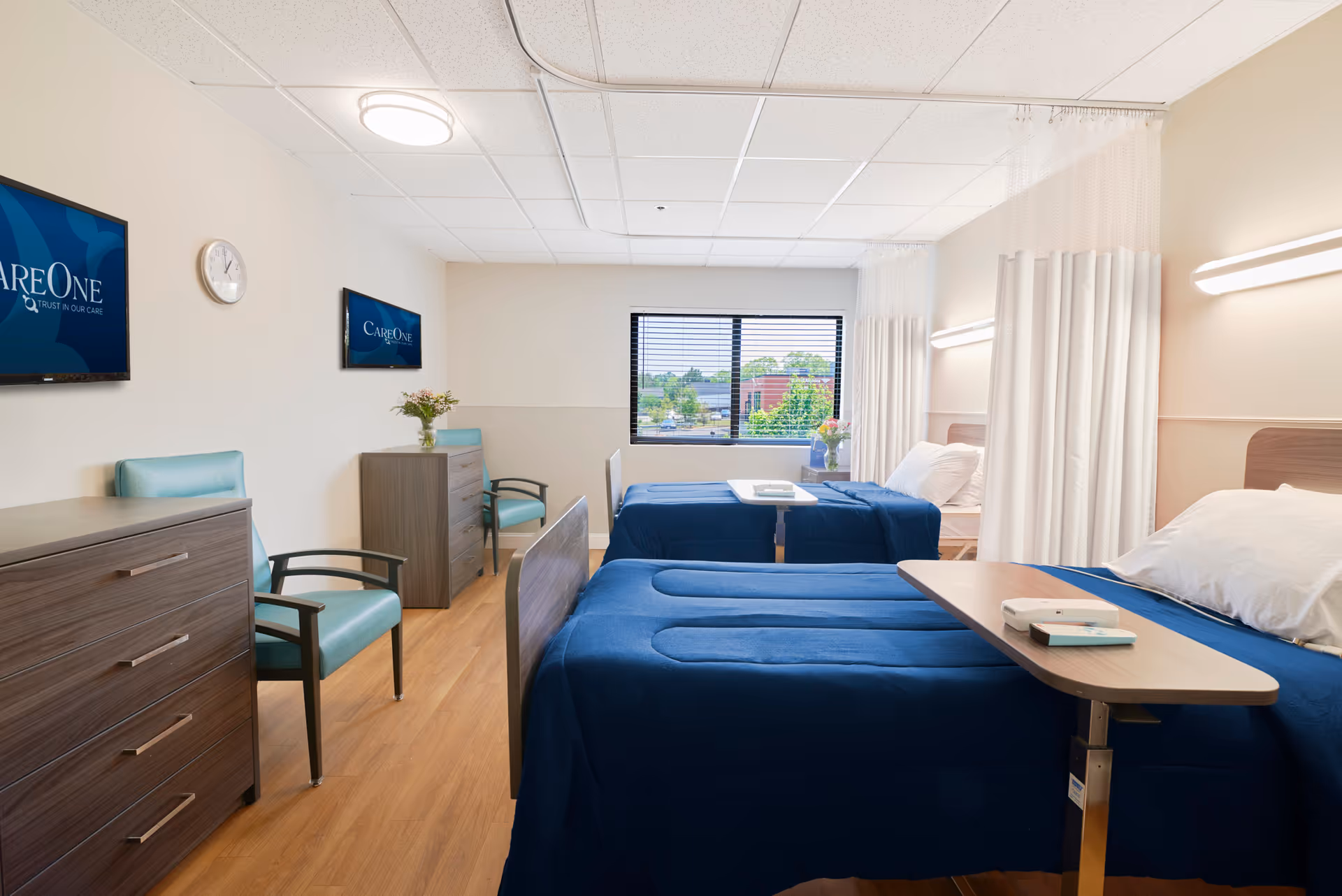 A bright and clean room with two hospital-style beds covered in blue bedding, each with a white pillow and an adjustable overbed table. The room has light-colored walls, wood flooring, two teal chairs, two dressers, and two wall-mounted televisions displaying the CareOne logo. A window at the far end shows a view of trees and buildings outside. Privacy curtains are partially drawn around each bed.