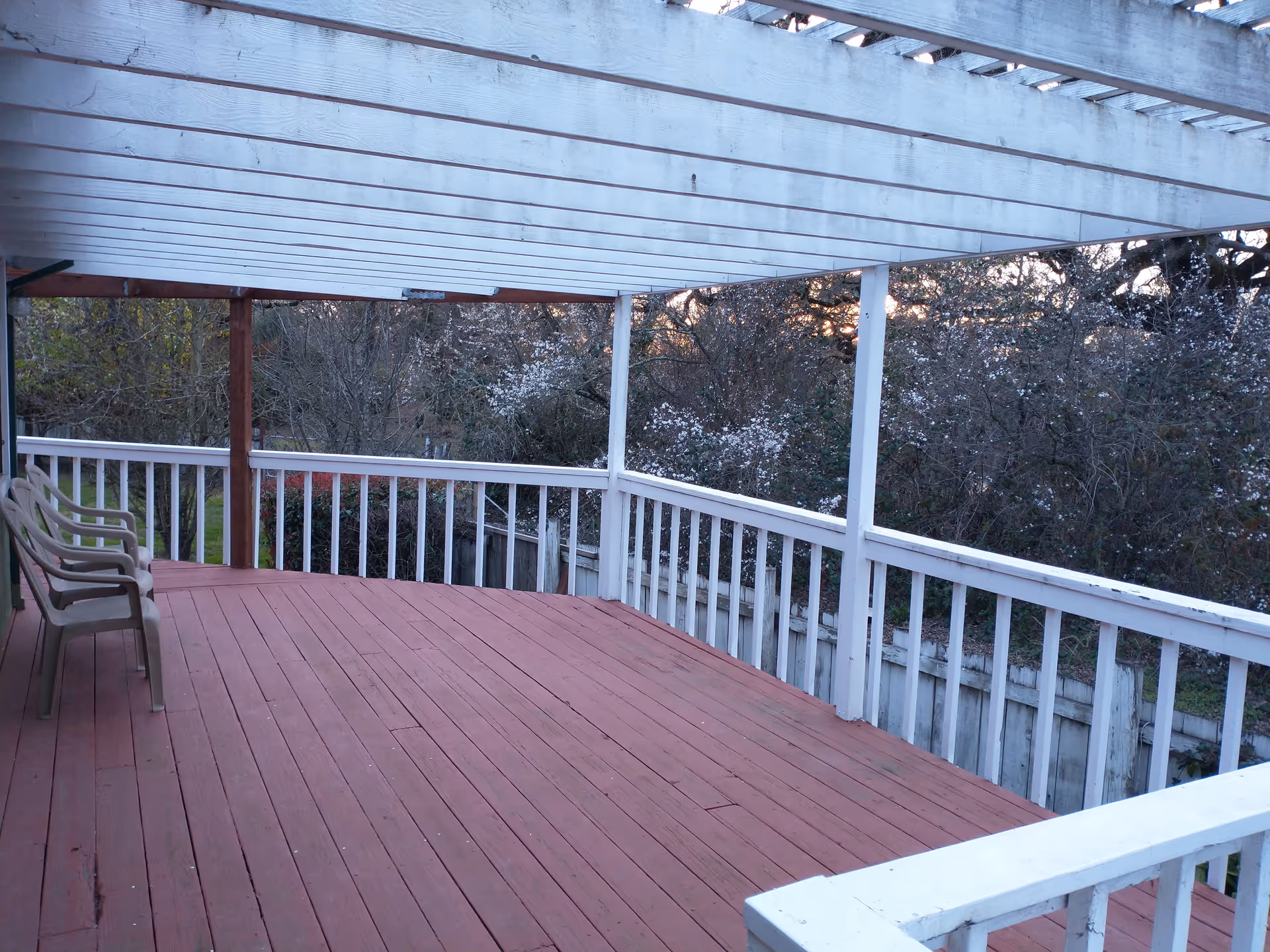 A covered outdoor wooden deck with a red floor and white railing. There are three plastic chairs lined up on the left side. Trees and bushes are visible beyond the railing.