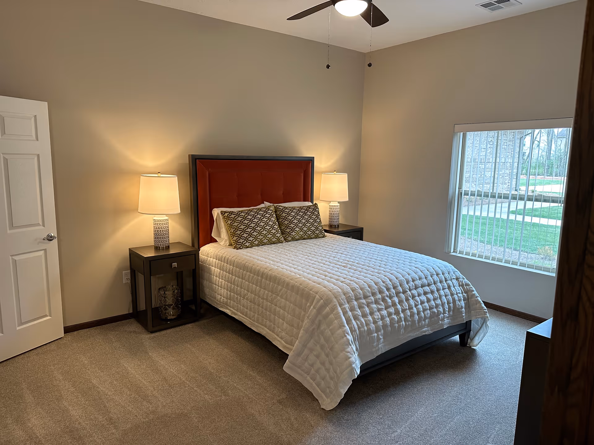 A neatly made bed with a red upholstered headboard and white quilted bedding in a bedroom. There are two patterned pillows on the bed and matching bedside tables with lamps on each side. A window with vertical blinds lets in natural light, and a ceiling fan with a light is visible above.