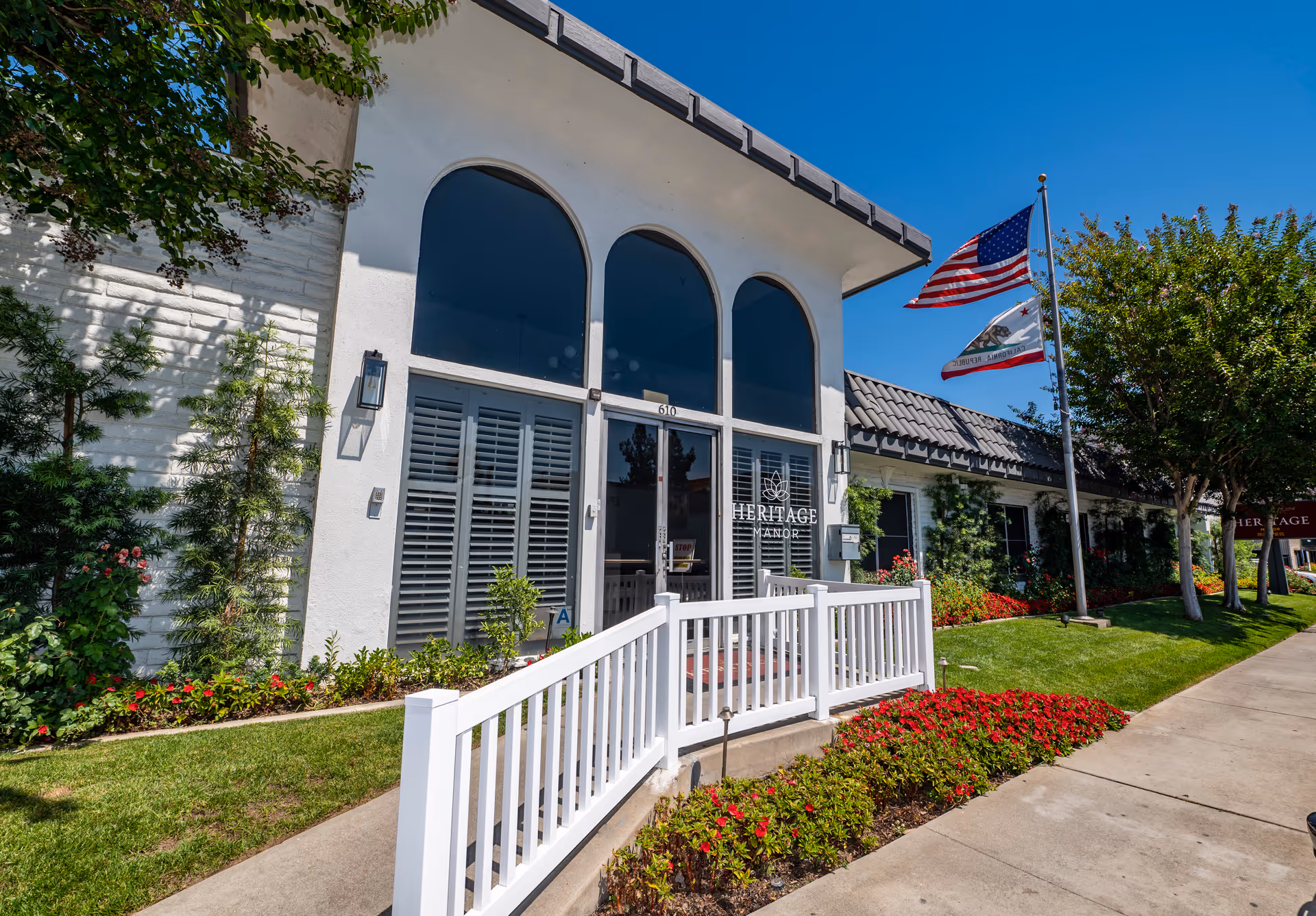 Entrance facade of Heritage Manor senior living facility with white railing, arched windows, American and California flags, and landscaped flower beds.