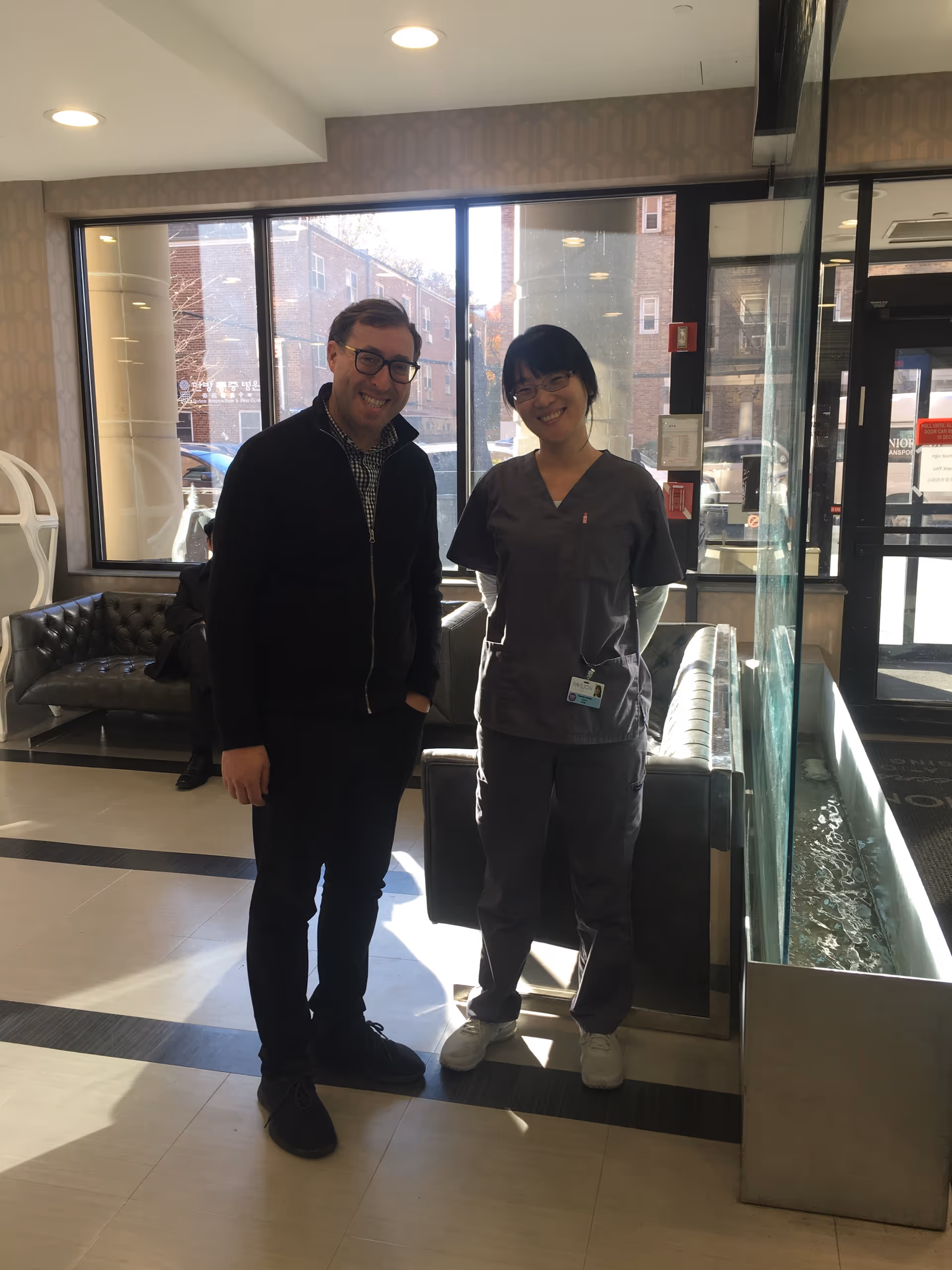 A man and a woman standing and smiling inside a well-lit lobby area with large windows. The woman is wearing gray medical scrubs and white shoes, while the man is dressed in dark casual clothing and glasses. Behind them are leather couches and a water feature along the right side.