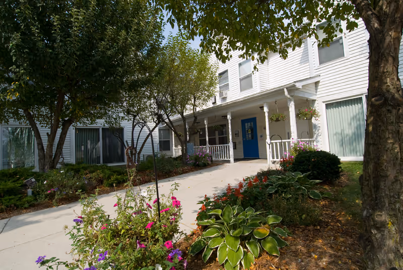 Exterior view of a white senior living facility building with a blue door entrance, surrounded by trees, shrubs, and colorful flowers along a concrete walkway.