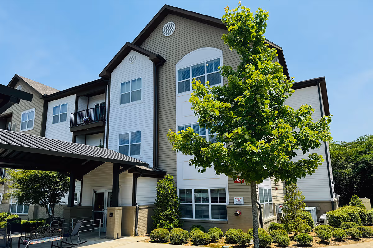 Exterior view of a multi-story senior living facility building with beige and white siding, several windows, a covered entrance, and landscaped greenery including a tree and bushes under a clear blue sky.