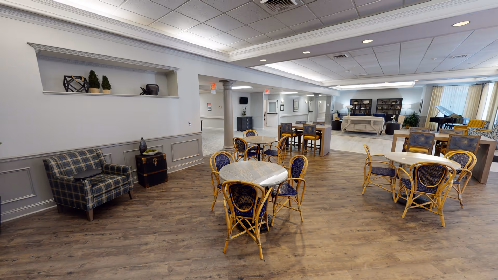 A spacious senior living common area with multiple round tables and wicker chairs arranged on a wood floor. In the background, there is a sitting area with sofas, bookshelves, and a grand piano near large windows with curtains. The walls are light-colored with decorative molding and columns, and the ceiling has recessed lighting.