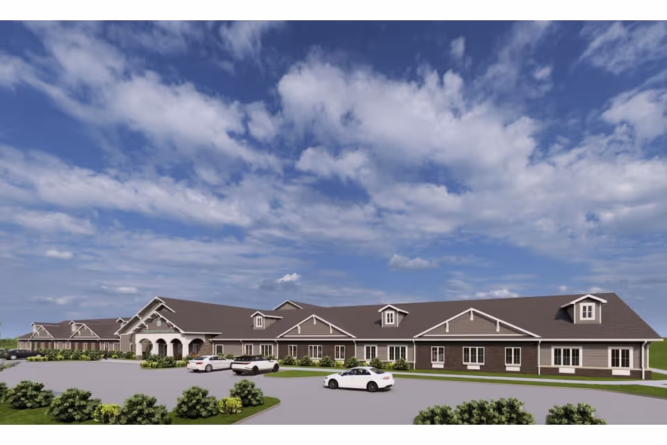Exterior view of a single-story senior living facility building with a large parking area in front, several cars parked, and a partly cloudy blue sky overhead.