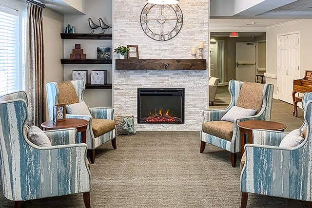 A cozy sitting area in a senior living facility with four upholstered armchairs arranged around two small round wooden tables. The chairs have a blue and beige striped pattern with beige cushions. A modern electric fireplace with a stone surround and a wooden mantel is centered on the wall, above which hangs a large decorative clock. Shelves with decorative items are on the left side, and a piano is visible on the right side in the background.