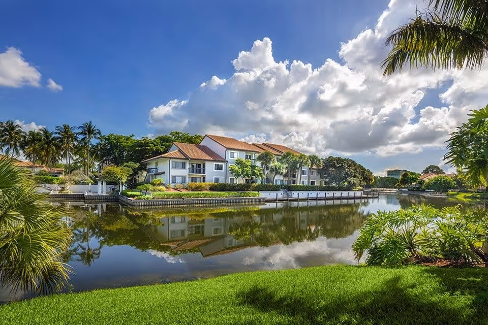 A serene outdoor view of a residential building with red-tiled roofs reflected in a calm body of water, surrounded by lush green grass, palm trees, and other tropical plants under a partly cloudy blue sky.