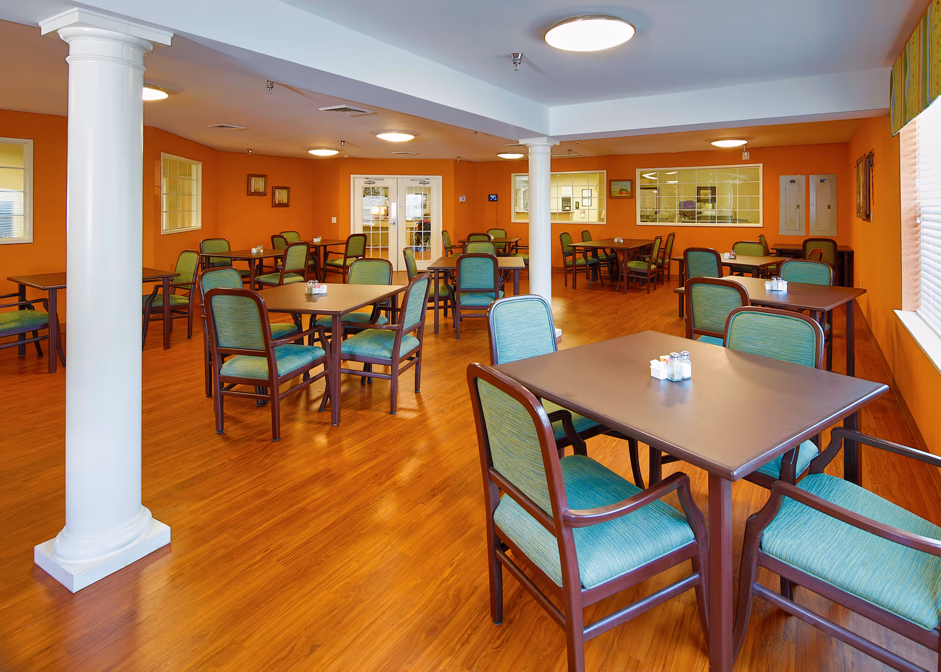 A spacious dining room with multiple square tables and green cushioned chairs arranged neatly on a wooden floor. The walls are painted orange, and there are white columns in the room. The room is well-lit with ceiling lights and natural light coming from a window on the right side. Salt and pepper shakers are placed on each table.